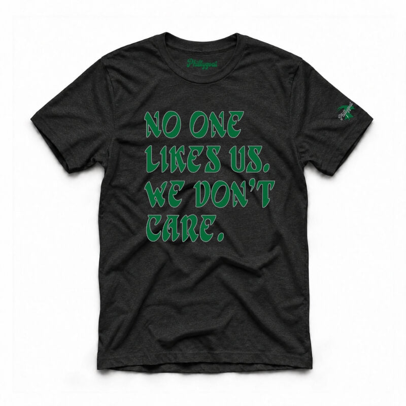 A dark charcoal gray heather t-shirt with the phrase 'NO ONE LIKES US, WE DON'T CARE.' printed in a stylized green font on the front.
