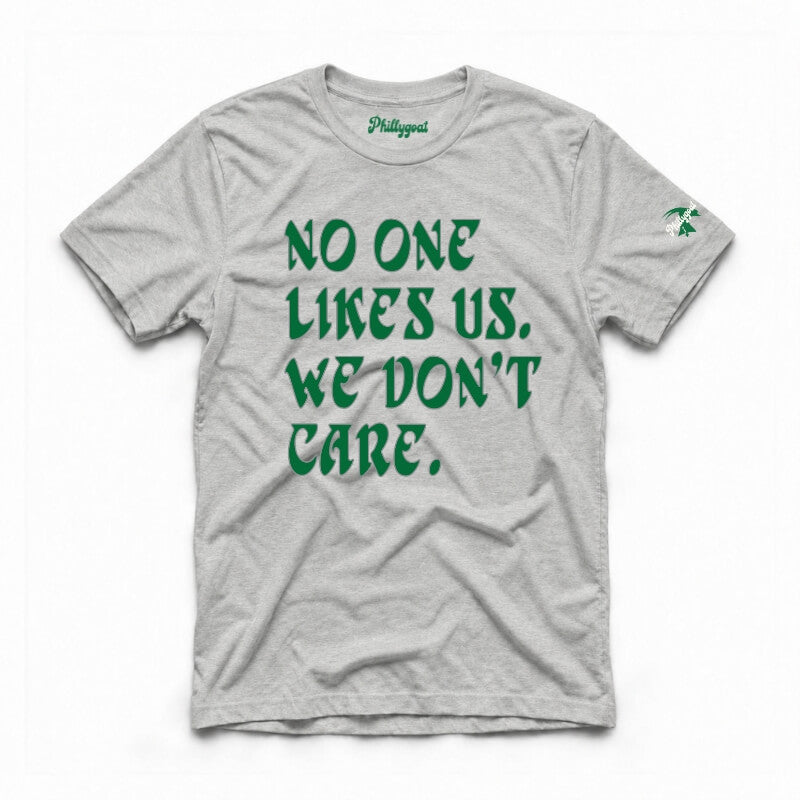 A light heather gray t-shirt with bold green text reading 'NO ONE LIKES US, WE DON'T CARE.' printed on the front.