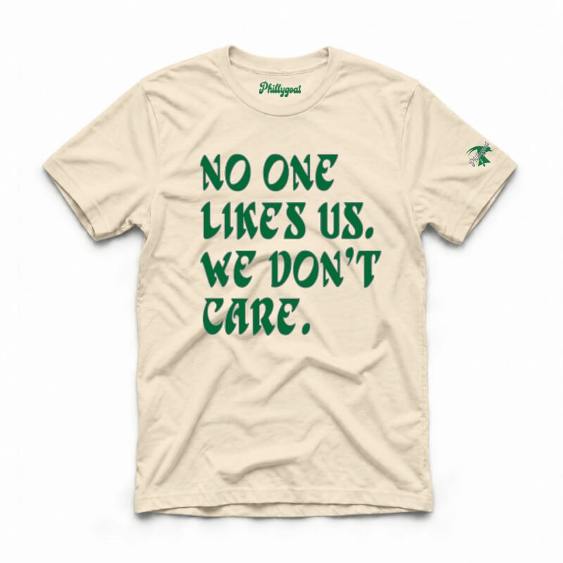 A beige crewneck t-shirt with green text reading 'NO ONE LIKES US, WE DON'T CARE.' printed on the front, featuring a small green graphic on the left sleeve and 'Philippist' printed inside the collar, shown on a plain white background.