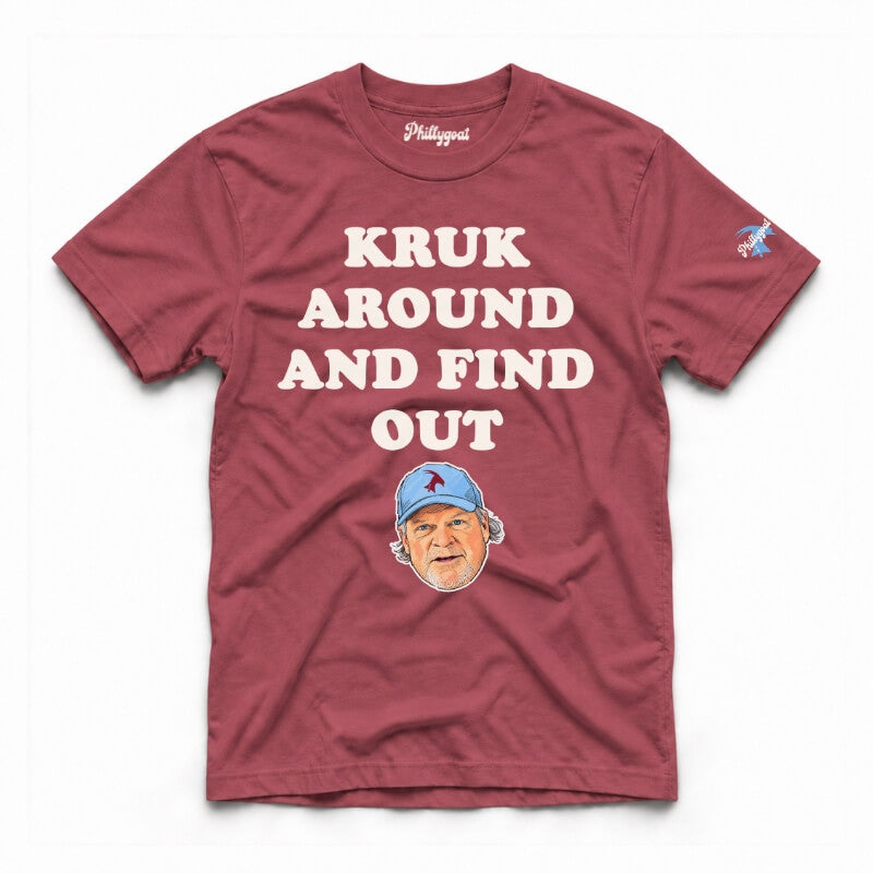 A maroon short-sleeve graphic t-shirt featuring the text 'KRUK AROUND AND FIND OUT' in cream block letters, a cartoon caricature of John Kruk wearing a blue cap, and a small blue logo on the right sleeve, displayed flat against a white background.