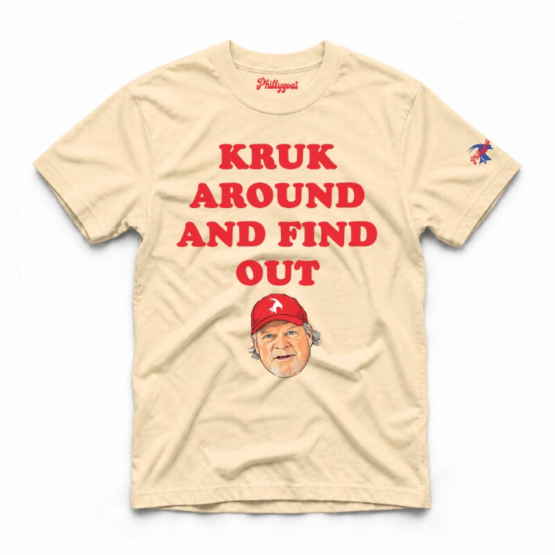 A ivory short-sleeve graphic t-shirt featuring the text 'KRUK AROUND AND FIND OUT' in cream block letters, a cartoon caricature of John Kruk wearing a blue cap, and a small blue logo on the right sleeve, displayed flat against a white background.