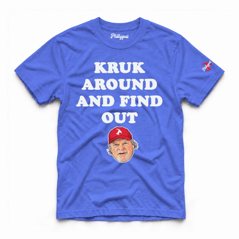 A royal blue short-sleeve graphic t-shirt featuring the text 'KRUK AROUND AND FIND OUT' in cream block letters, a cartoon caricature of John Kruk wearing a blue cap, and a small blue logo on the right sleeve, displayed flat against a white background.