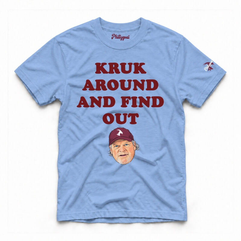 A washed denim short-sleeve graphic t-shirt featuring the text 'KRUK AROUND AND FIND OUT' in cream block letters, a cartoon caricature of John Kruk wearing a blue cap, and a small blue logo on the right sleeve, displayed flat against a white background.